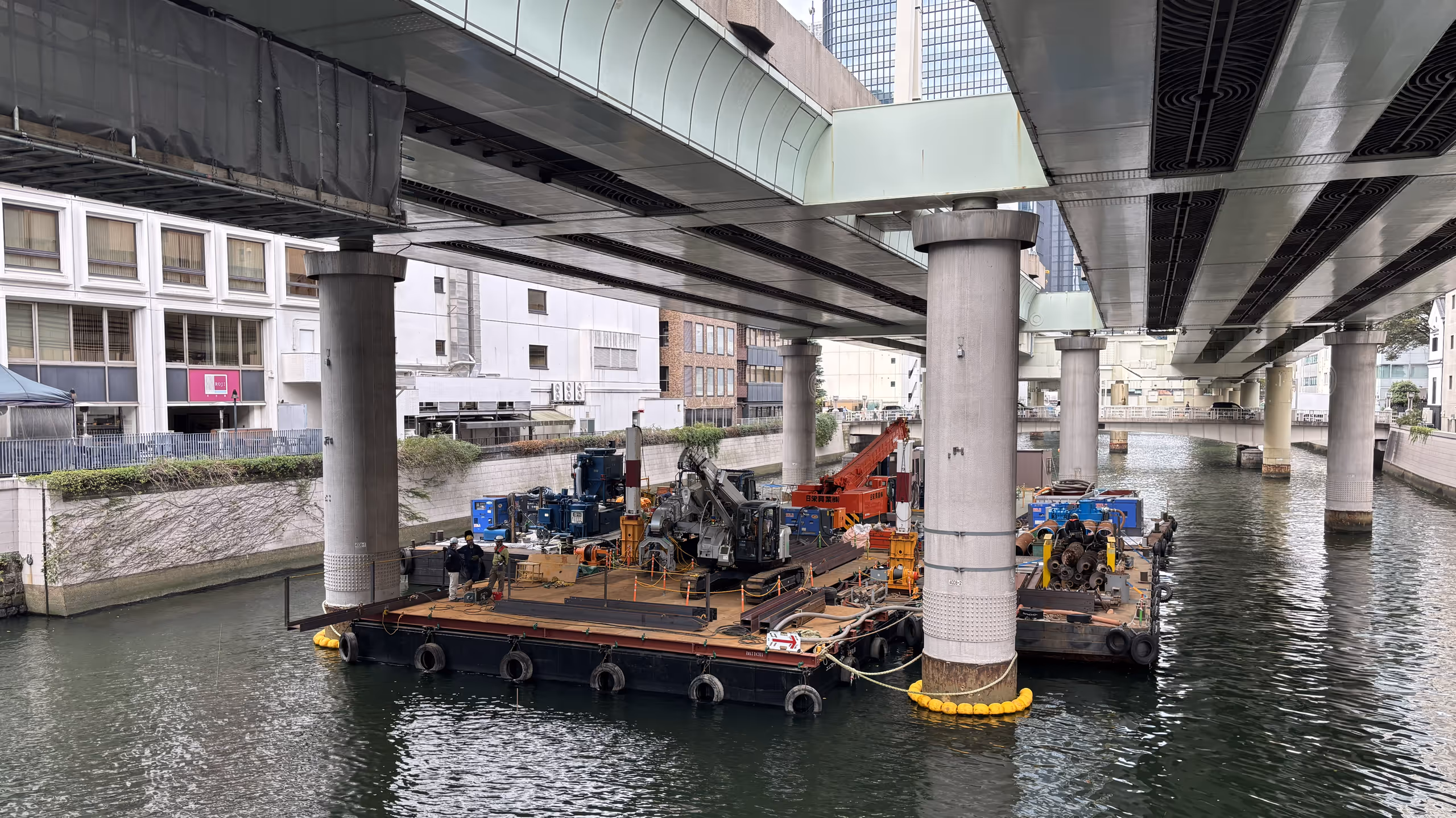 高速道路の下を通る川に、広い足場が浮いており、何台かの重機が作業をしている。